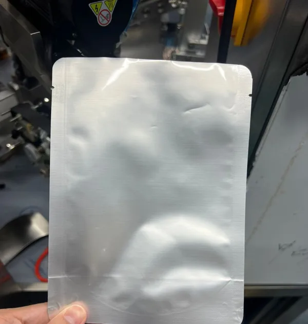 Hand holding a plain unprinted foil retort pouch in front of a premade pouch machine